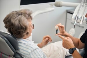 An older woman sits in a dental chair while a dentist wearing a black shirt and watch shows her a dental implant model. Dental equipment and a monitor are visible in the background.